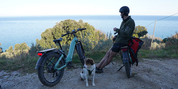 Zwei Himiway D5 Fatbikes in Blau und Schwarz auf einem Berg mit Meerblick, ein Mann lehnt am schwarzen Fatbike, neben ihm sitzt ein Hund.